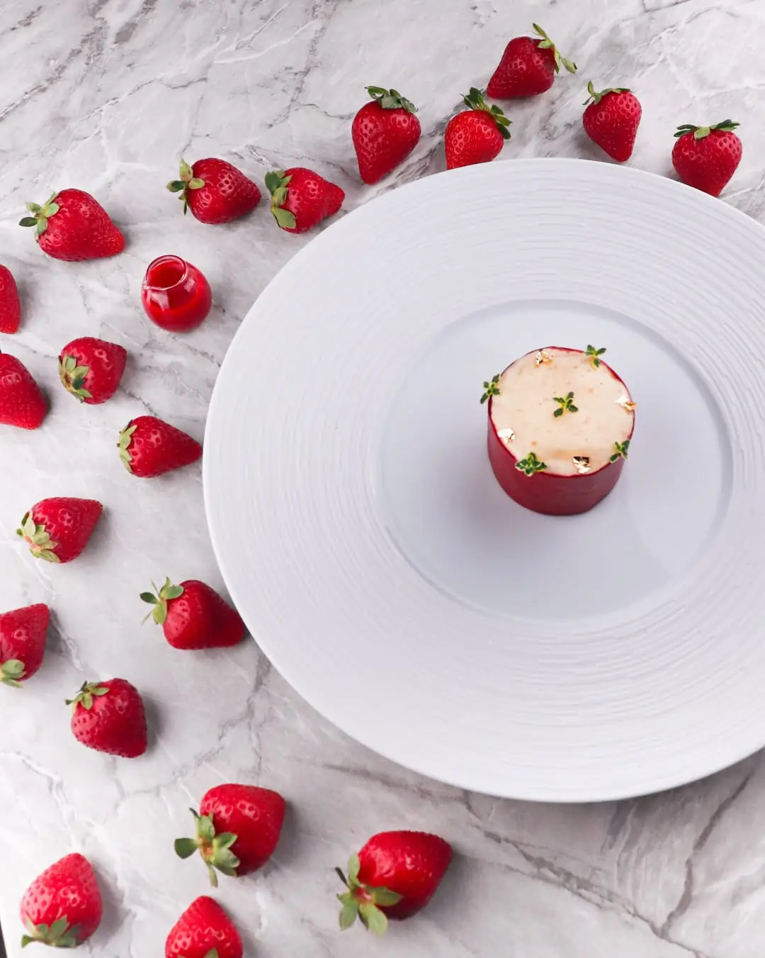 Small red mousse cake topped with gold leaf on a white plate ringed with fresh strawberries
