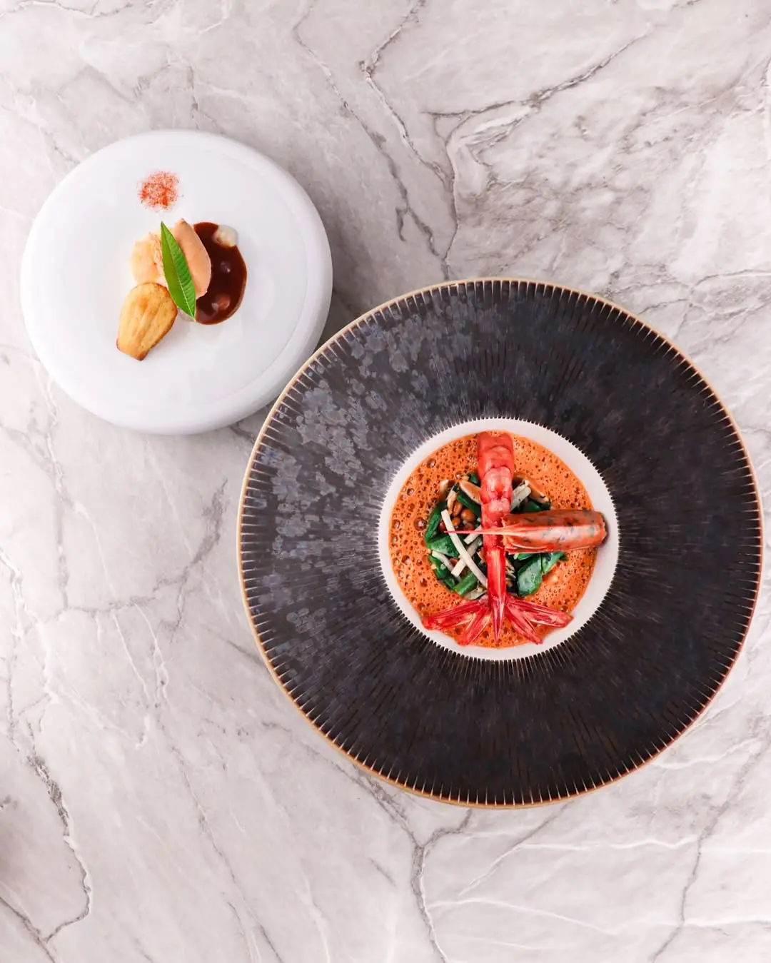 Whole grilled prawn over greens in a red bisque sauce next to a small madeleine and sauce plate