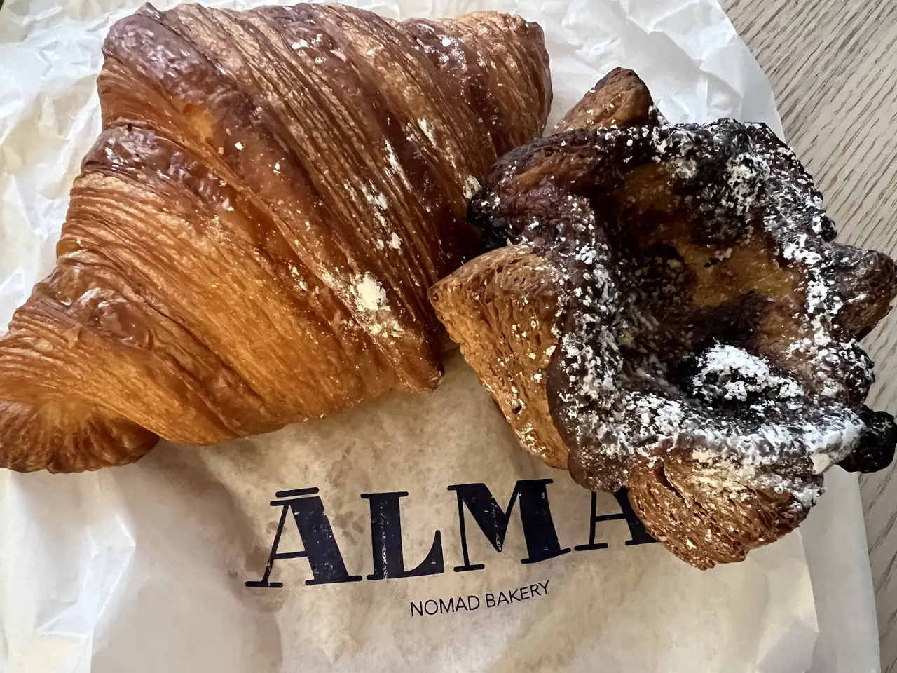 A plain croissant and a chocolate pastry dusted with powdered sugar on branded bakery paper