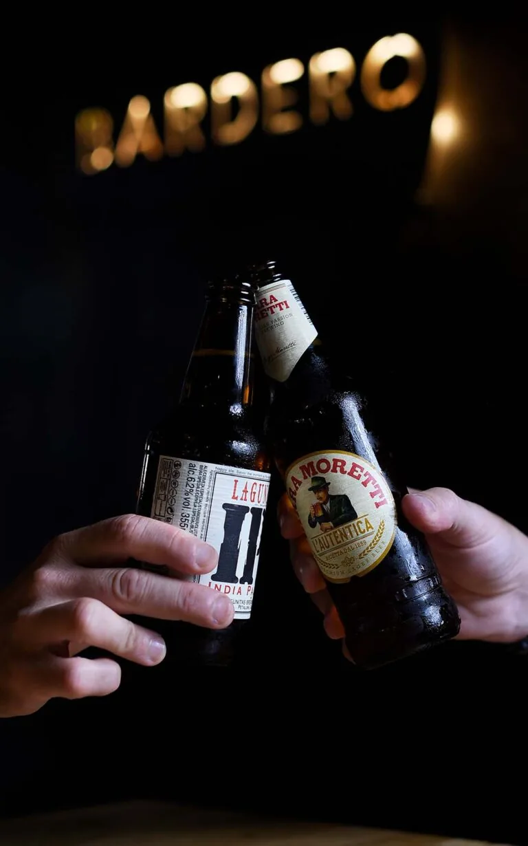 Two hands clinking a Lagunitas IPA and a Birra Moretti bottle beneath the illuminated BARDERO letters on a dark wall