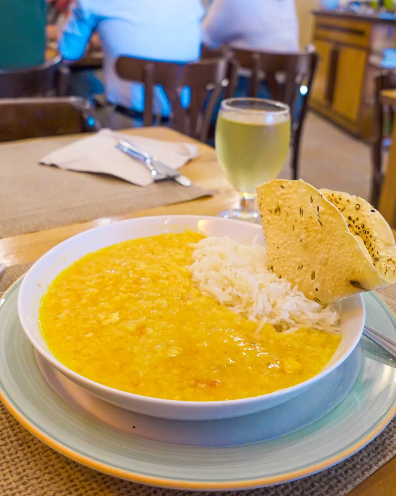 Bowl of yellow lentil dal with a scoop of white rice and a crisp papadum, served with a small glass of lassi, at Amaltea