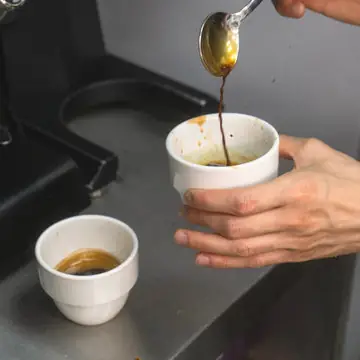 Espresso being spooned and poured between two white cups at the machine