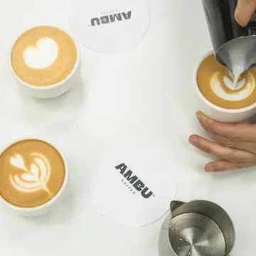 Three latte-art coffees on AMBU-branded coasters with milk being poured into one