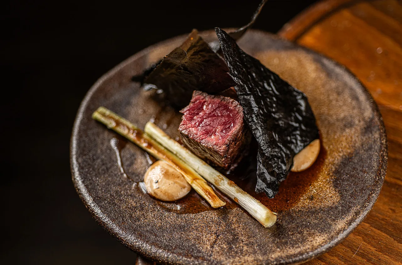 A cube of rare grilled beef on a rough dark ceramic plate with charred sheets of seaweed, two grilled spring onions and a quenelle of pale sauce
