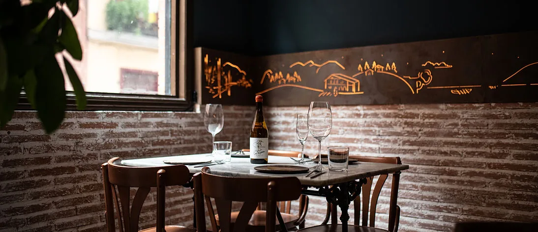A marble table laid for two beside a window, with a bottle of white wine and glasses, bentwood chairs, exposed brick and a copper wall panel etched with a landscape scene