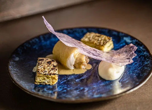 A rolled fillet of white fish in a pale sauce on a blue glazed plate, with a thin violet crisp, browned custard squares and a spoonful of foam