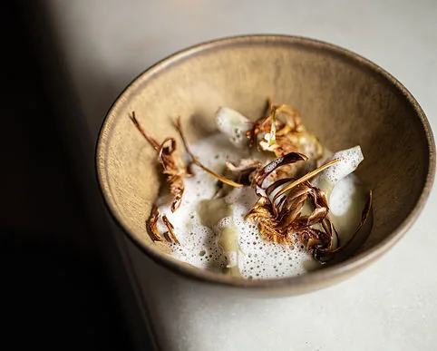 A shallow stoneware bowl of roasted shallot petals and white vegetable in a frothed pale broth, seen from above on a grey surface