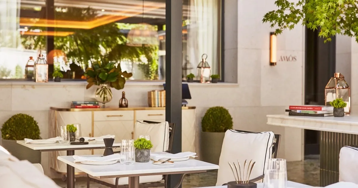 Outdoor tables set with glasses and napkins beside a sideboard with books and plants at the restaurant entrance