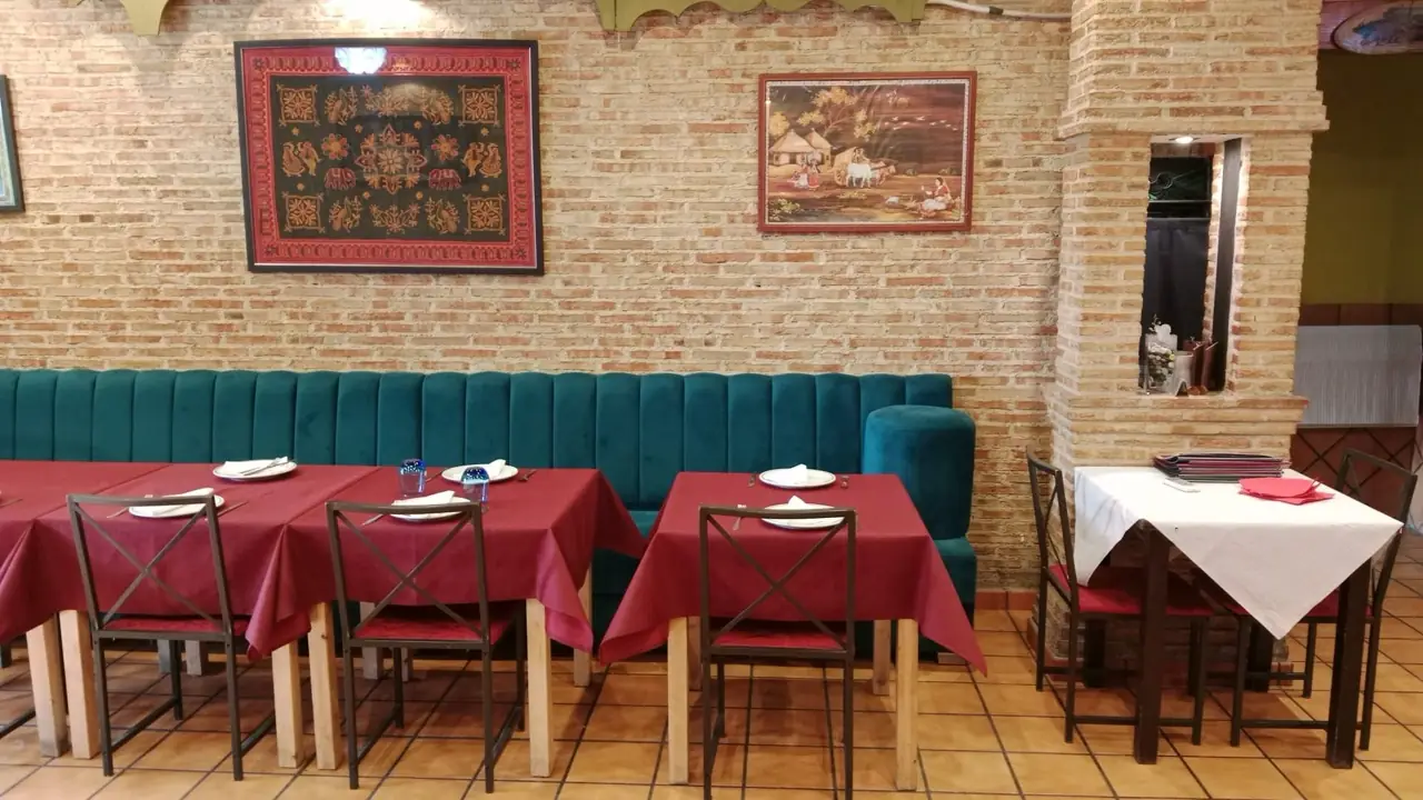 Indian restaurant dining room, tables with maroon cloths and set places against a brick wall hung with Indian tapestry art and a teal banquette