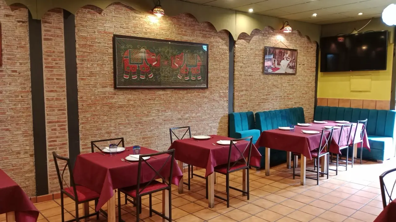 Dining room with maroon-clothed tables, teal banquette and a framed red elephant tapestry on the brick wall beside a wall-mounted TV
