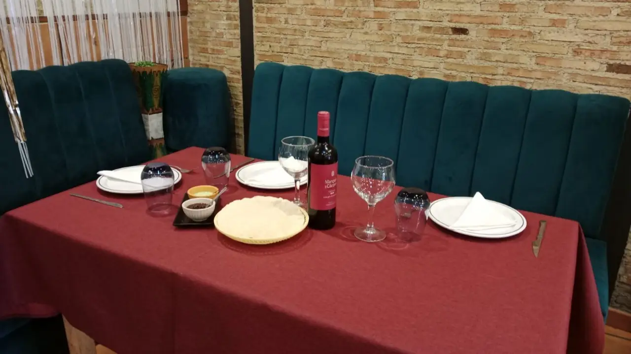Set table with a basket of poppadoms, small bowls of chutney, a bottle of Marques de Caceres red wine and wine glasses on a maroon cloth by a teal banquette