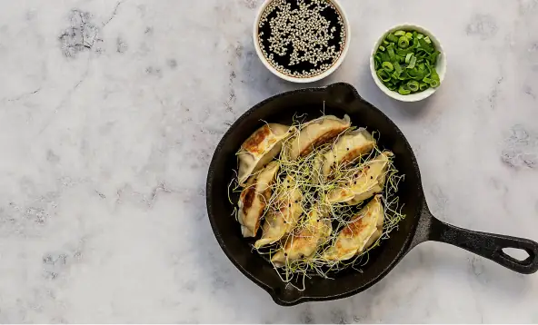 Pan-fried gyoza dumplings in a cast-iron skillet strewn with microgreens, with bowls of sesame and sliced scallion alongside