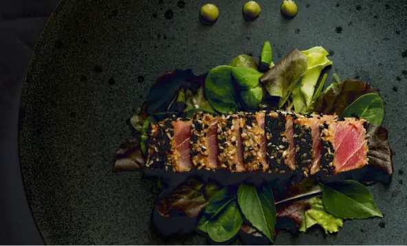 Sliced sesame-crusted seared tuna tataki fanned over mixed salad leaves on a dark plate with dots of sauce