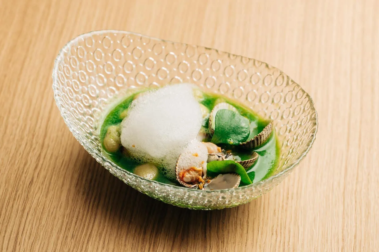 A dish with a white foam in a green broth in a textured glass bowl