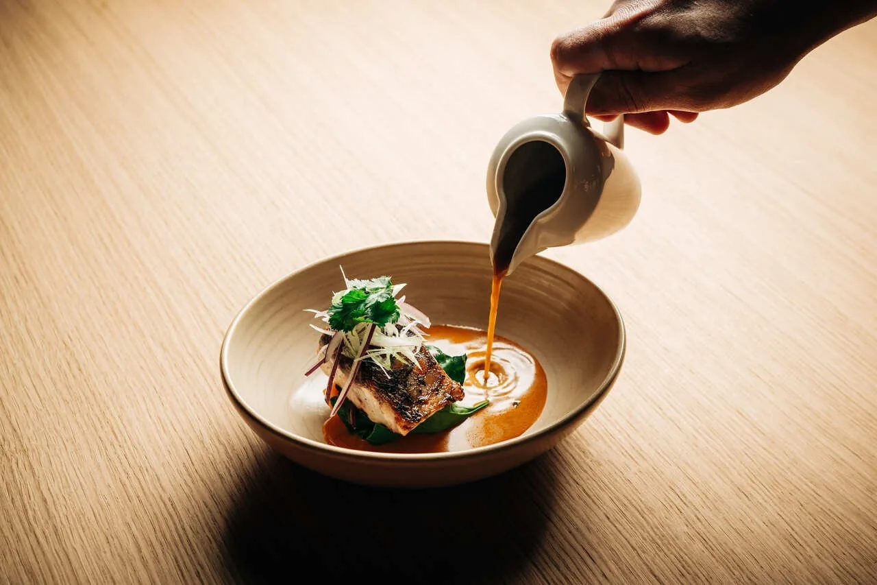 A sauce being poured into a bowl of fish with green vegetables
