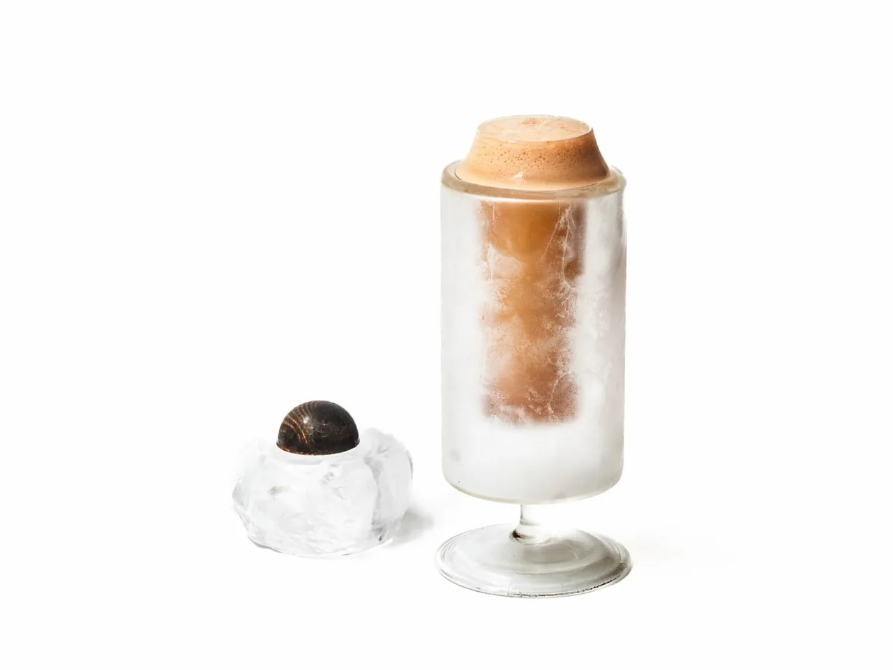 A cocktail glass with a frozen foam topping and a dark ball of ice on a white background
