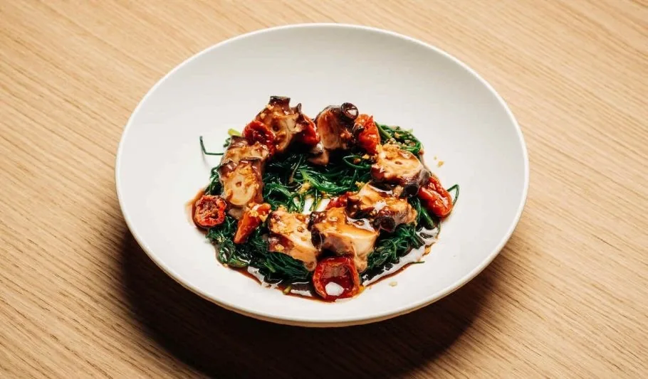 A white bowl of dark glazed pieces with greens