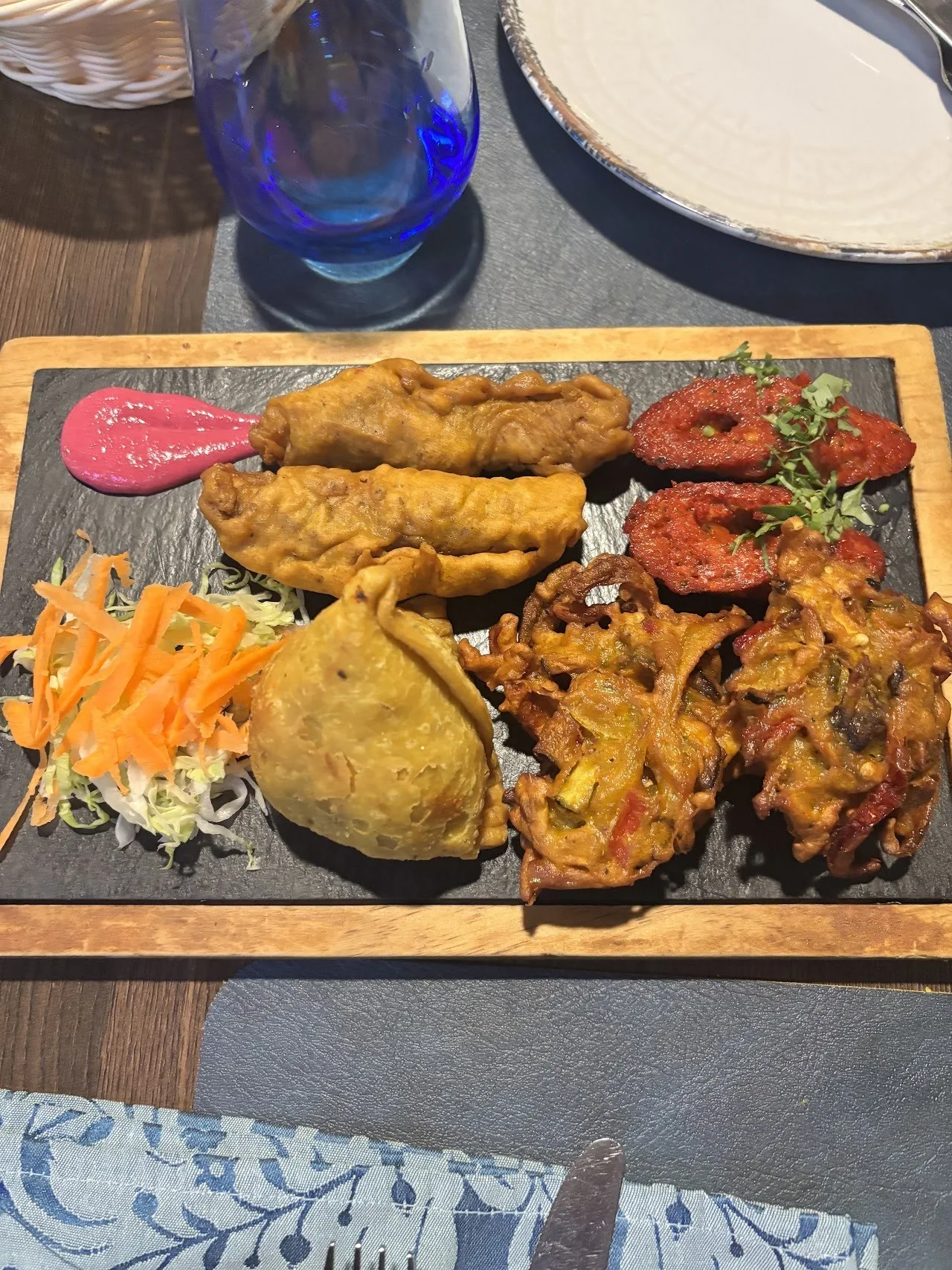 A slate board with fried fritters, a pastry and grilled meat pieces served with sliced carrot