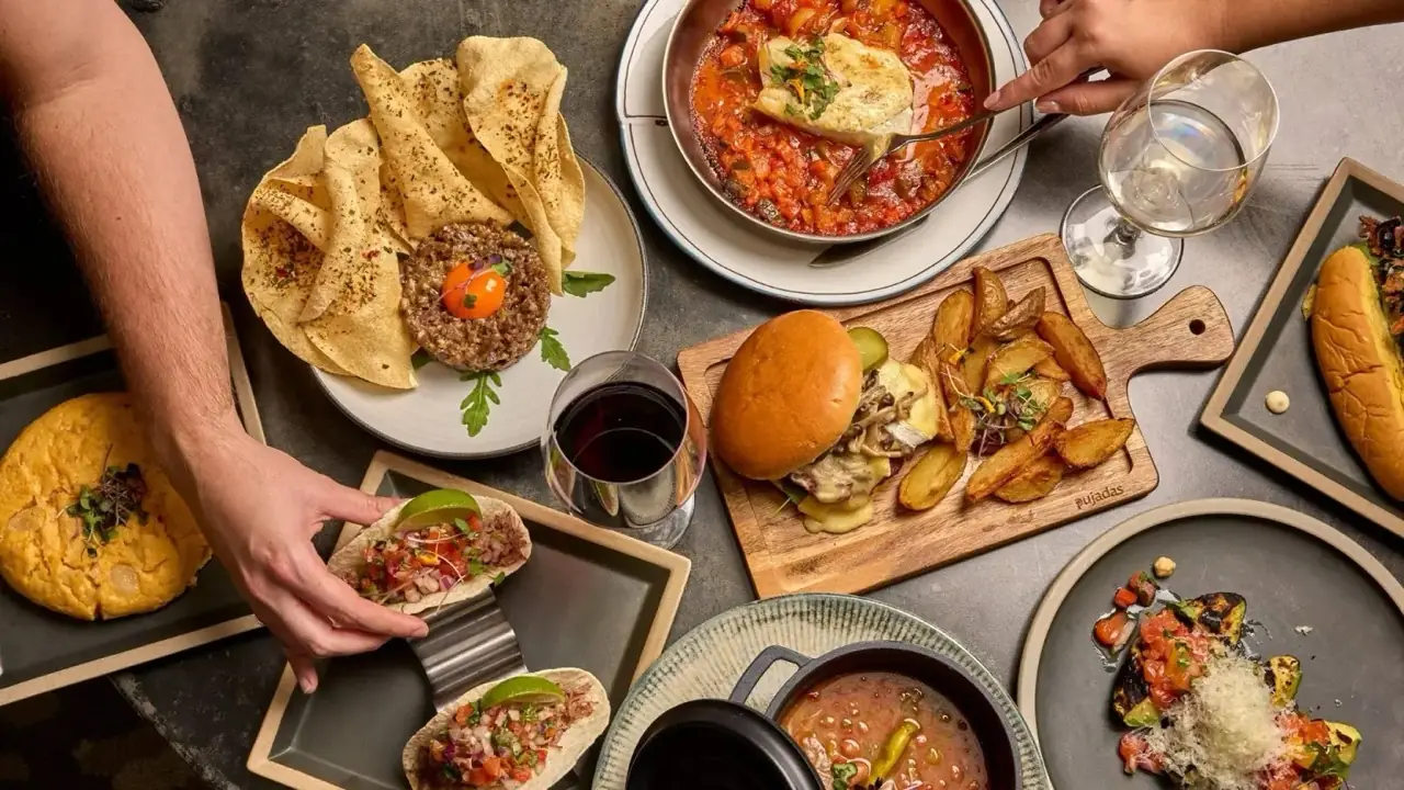 Overhead spread of shared dishes on a grey table: cod in tomato sauce, steak tartare with poppadoms, a burger with potato wedges, tacos, gazpacho and red and white wine