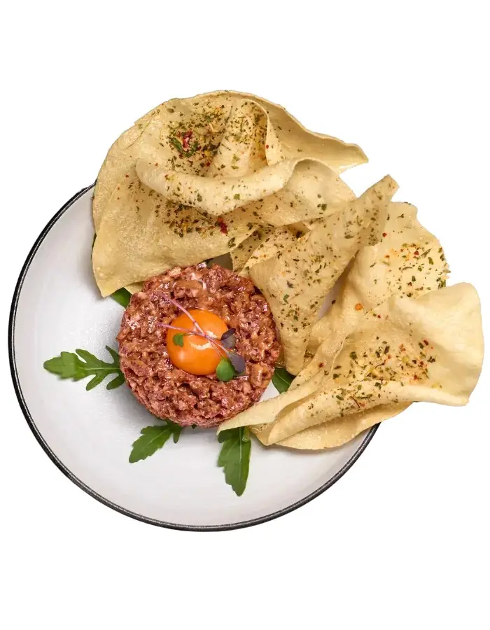 Steak tartare round topped with an orange yolk garnish, surrounded by seasoned poppadoms and rocket on a white plate