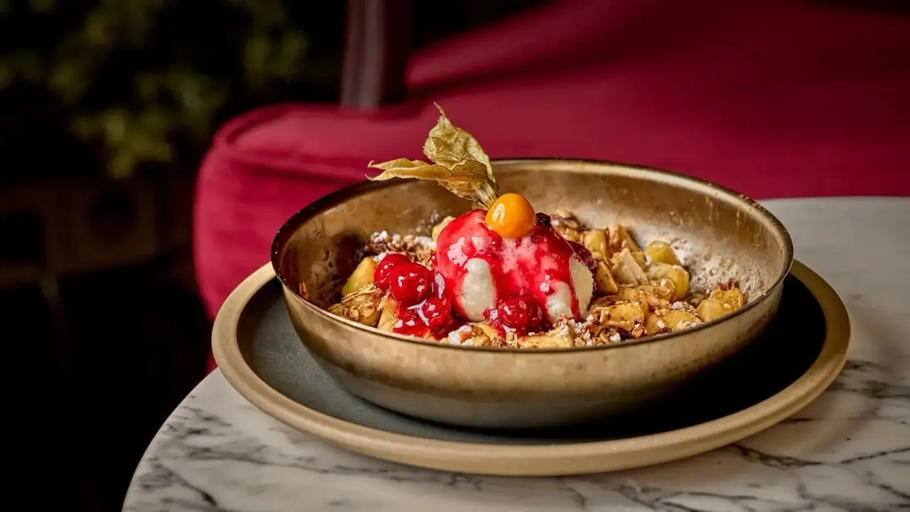 Apple crumble dessert in a brass bowl topped with ice cream, red berry sauce and a physalis, on a marble table