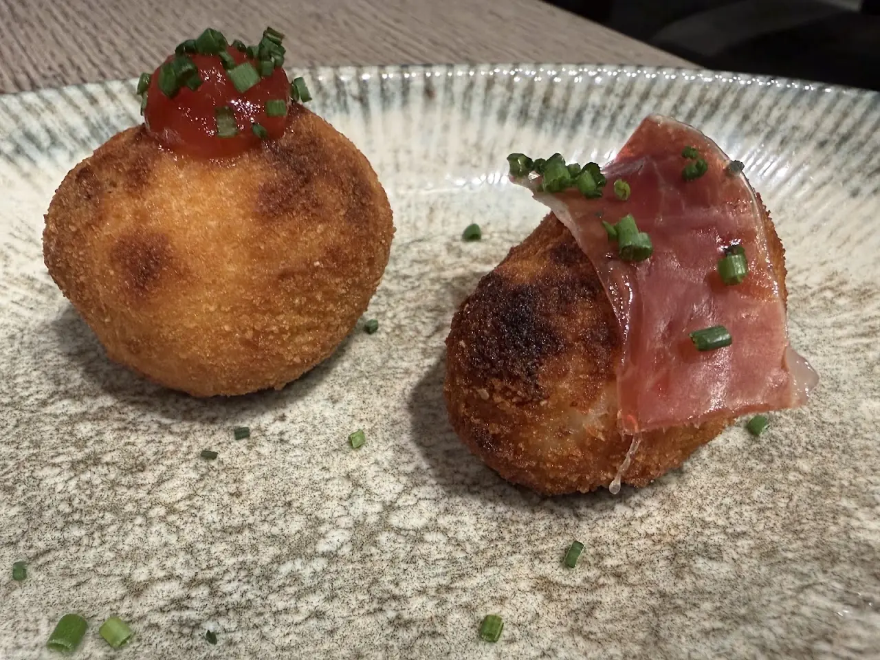 Two fried croquettes, one topped with cured ham and chives, on a textured plate.