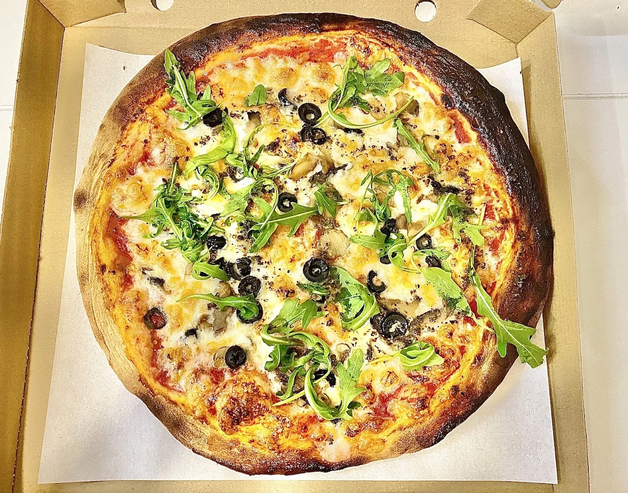 A pizza topped with arugula, black olives, and cheese in a takeout box