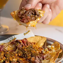 Hand holding a loaded tortilla chip with meat and herbs above a plate of nachos