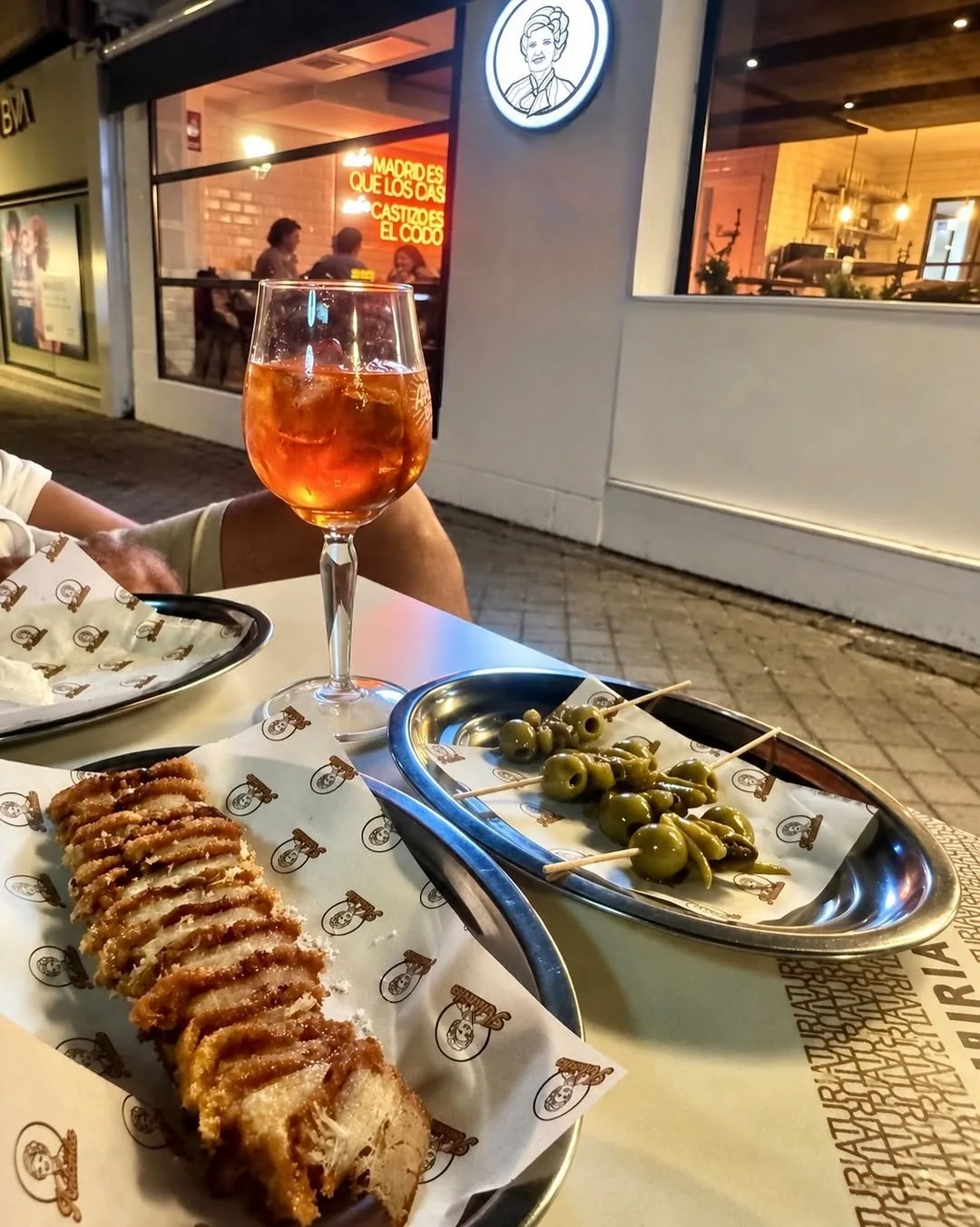 Sliced roast pork and green olives on a table beside a glass of orange spritz