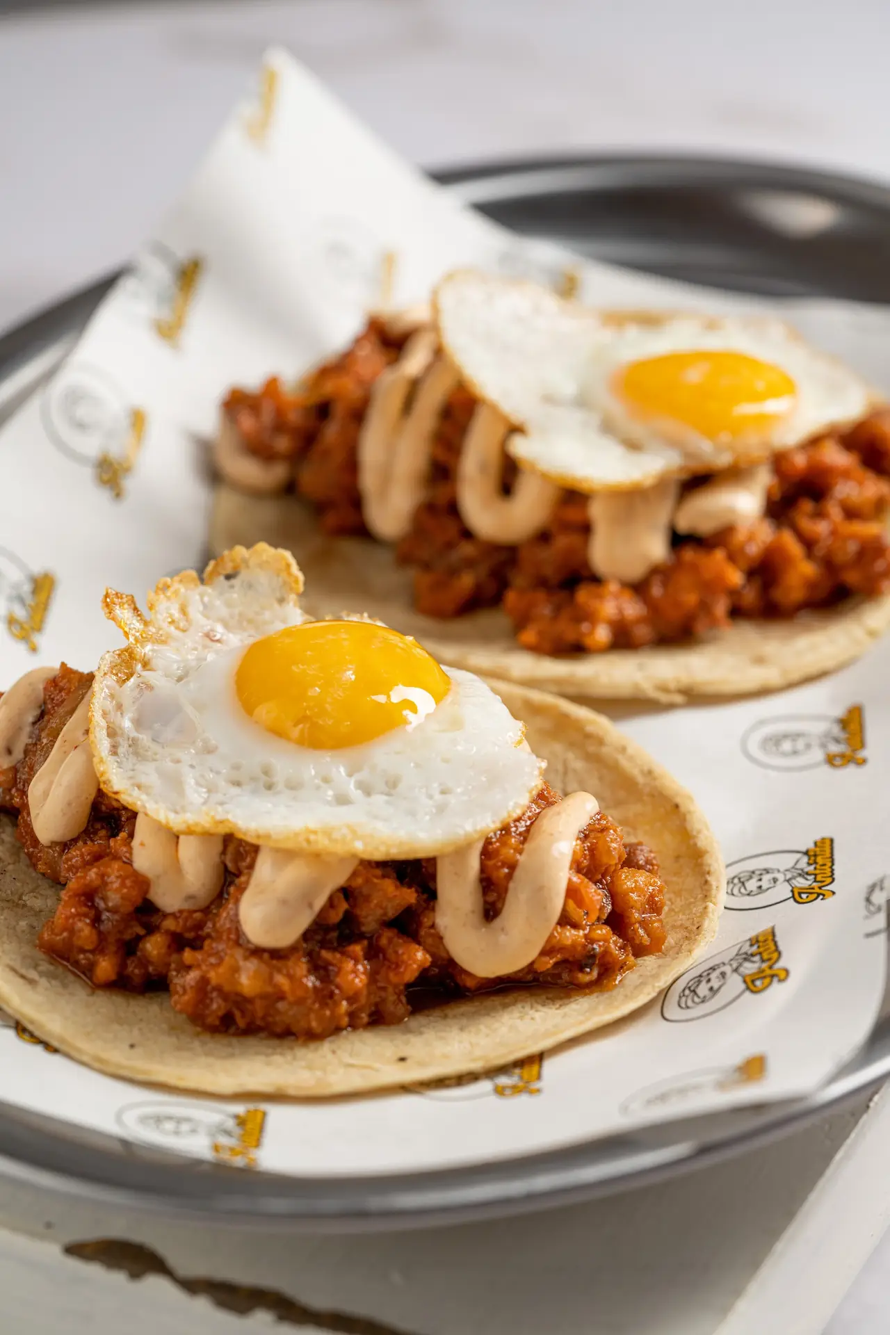 Two corn tacos topped with a spiced meat filling, sauce and a fried egg each, on branded paper