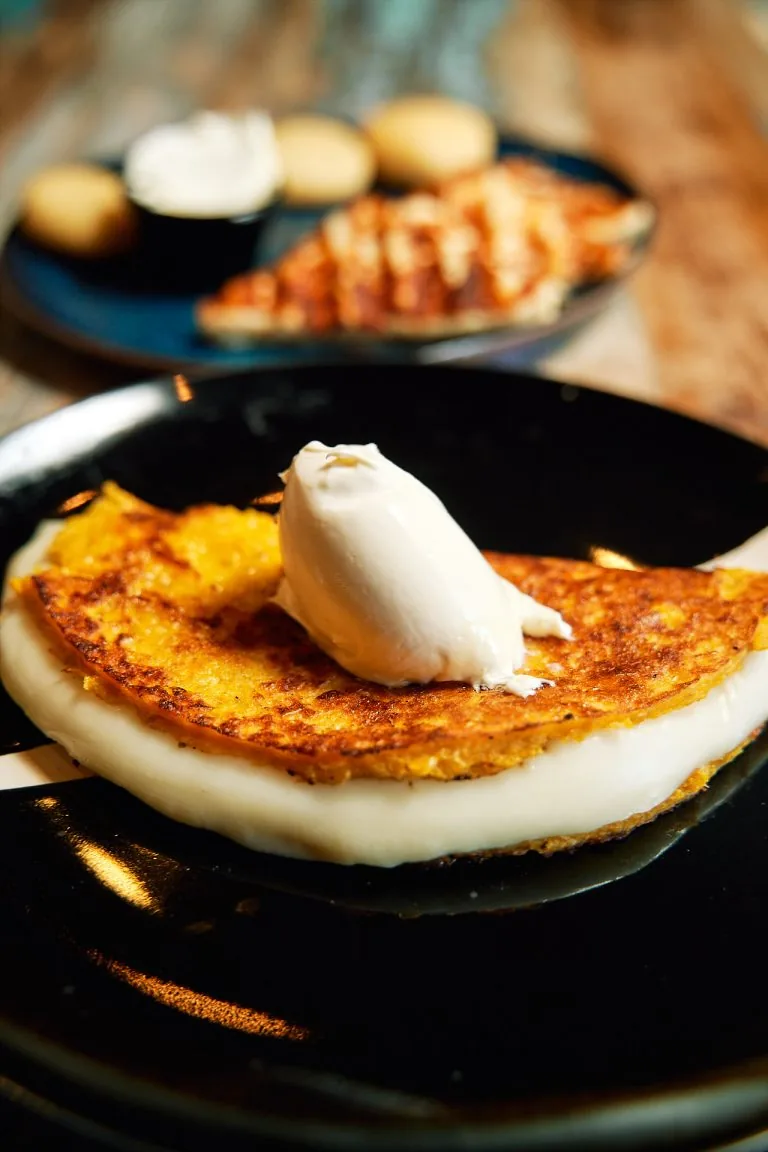 A golden corn cachapa folded over melting white cheese on a black plate, topped with a quenelle of cream, with a second plate of arepas and a cheese-crusted portion behind