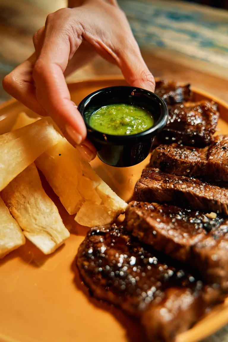 A hand lowering a small black pot of bright green sauce onto a terracotta plate of sliced grilled beef and fried yuca batons