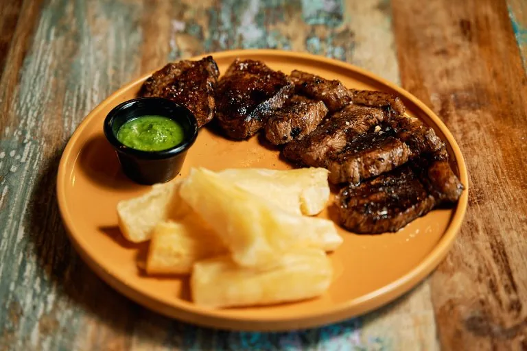 Sliced grilled beef with fried yuca batons and a pot of green sauce on a terracotta plate, on a distressed blue and brown wooden table