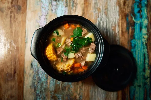 A black lidded pot of beef and vegetable soup with corn on the cob, potato, carrot and coriander, the lid resting beside it on a painted wooden table
