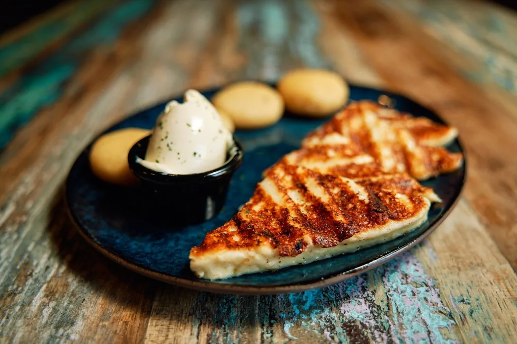 A grill-marked folded cheese arepa on a dark blue plate with small yellow dough balls and a pot of pale cream sauce