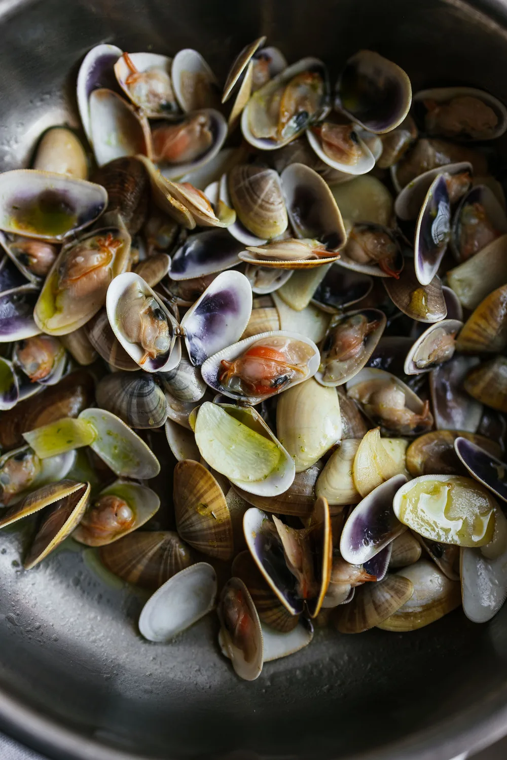 A steel pan filled with cooked clams, shells opened to show the meat, glossy with olive oil and flecks of green parsley