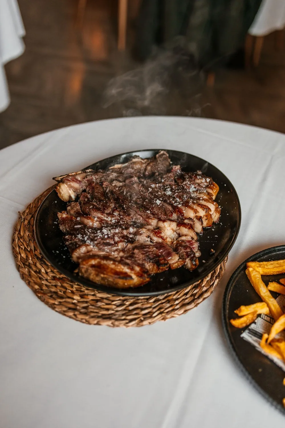 A grilled cut of meat on a round woven mat on a white tablecloth, with fries at the edge