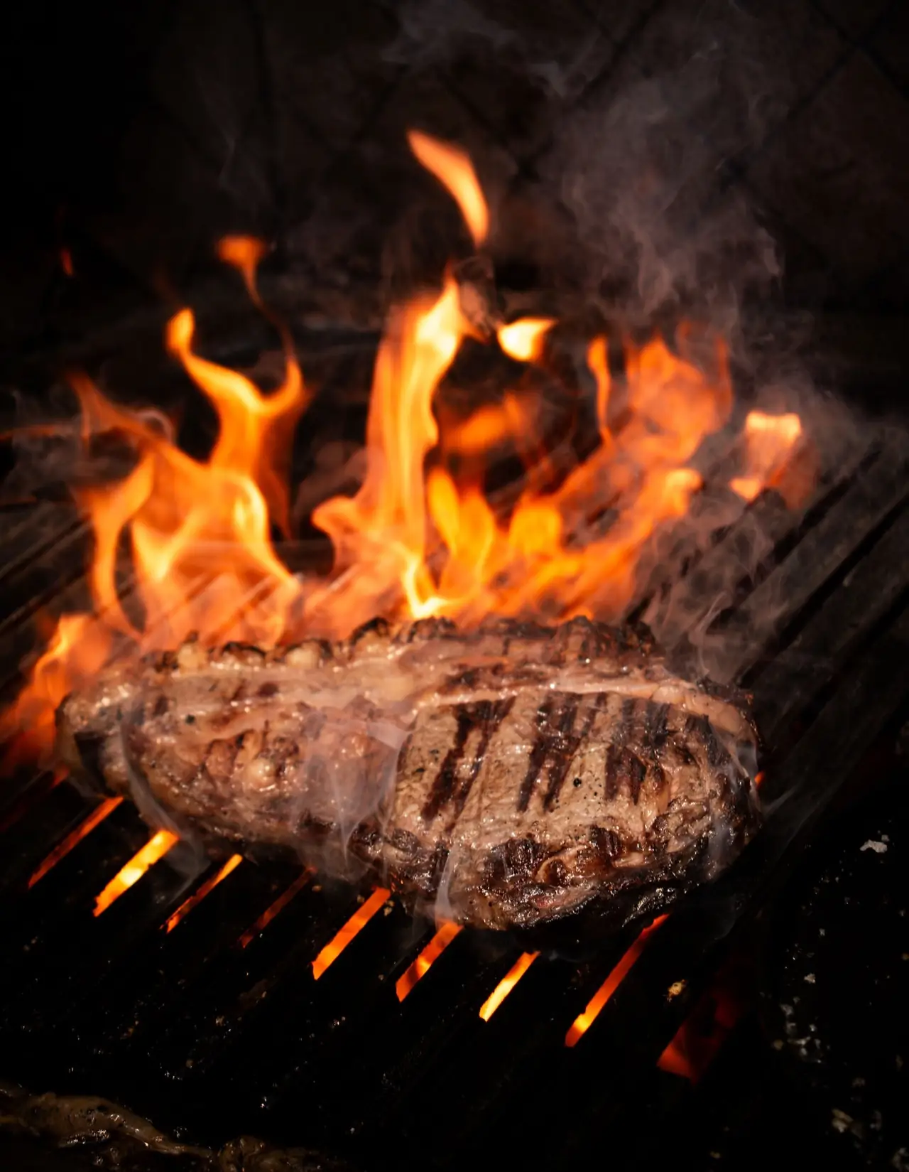 Thick steak with charred grill marks engulfed in open flames on a barbecue grill, smoke rising.