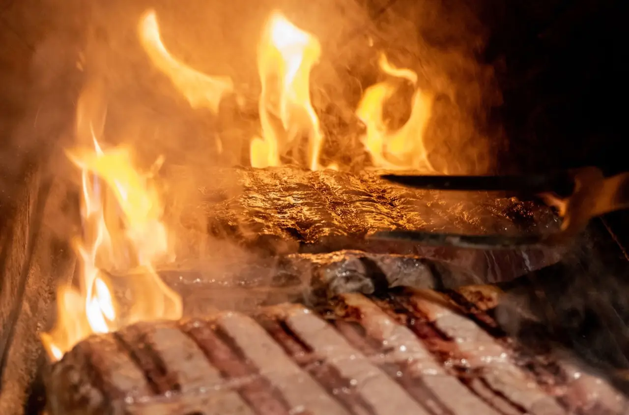 Hand using long tongs to turn a large cut of meat over an open flame inside a wood-fired grill.