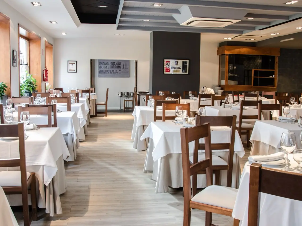 Restaurant dining room with white-clothed tables set with wine glasses and wooden chairs, dark accent wall and stone floor.
