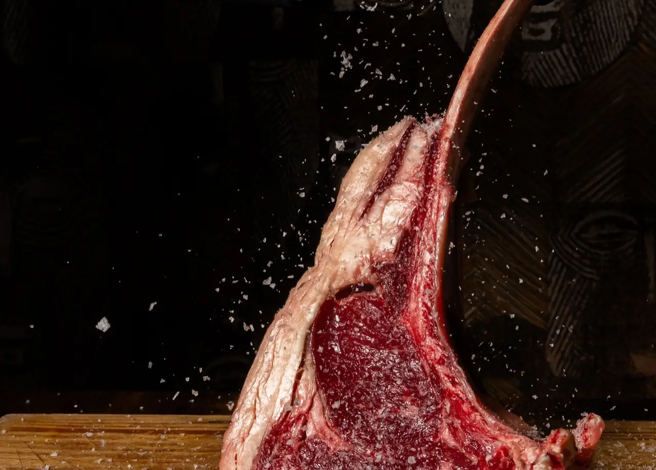 Raw bone-in tomahawk steak on a dark background with coarse salt crystals falling over it.