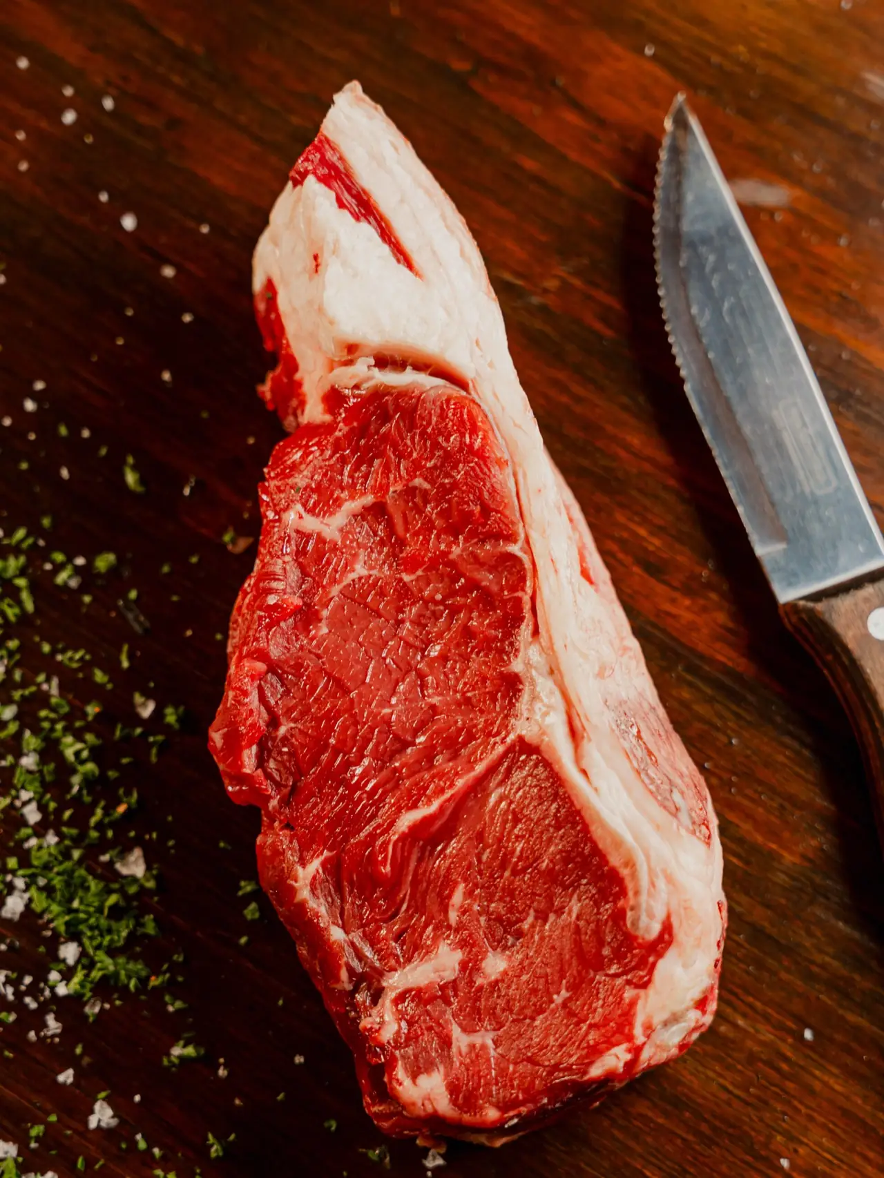 Raw marbled steak on a dark wood board next to a serrated knife, scattered with coarse salt and chopped herbs.