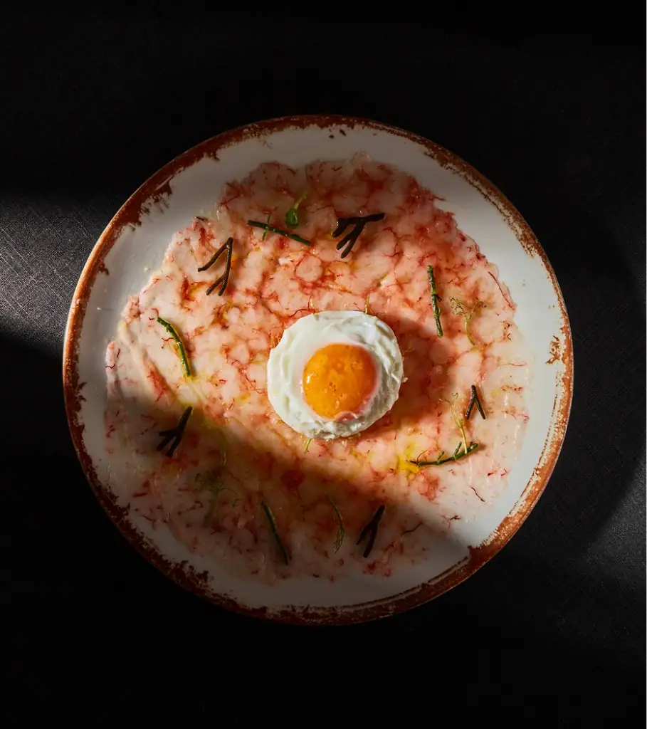 Round plate of thin-sliced pink carpaccio-style dish topped with a fried quail egg with runny yolk, garnished with herbs, overhead view.