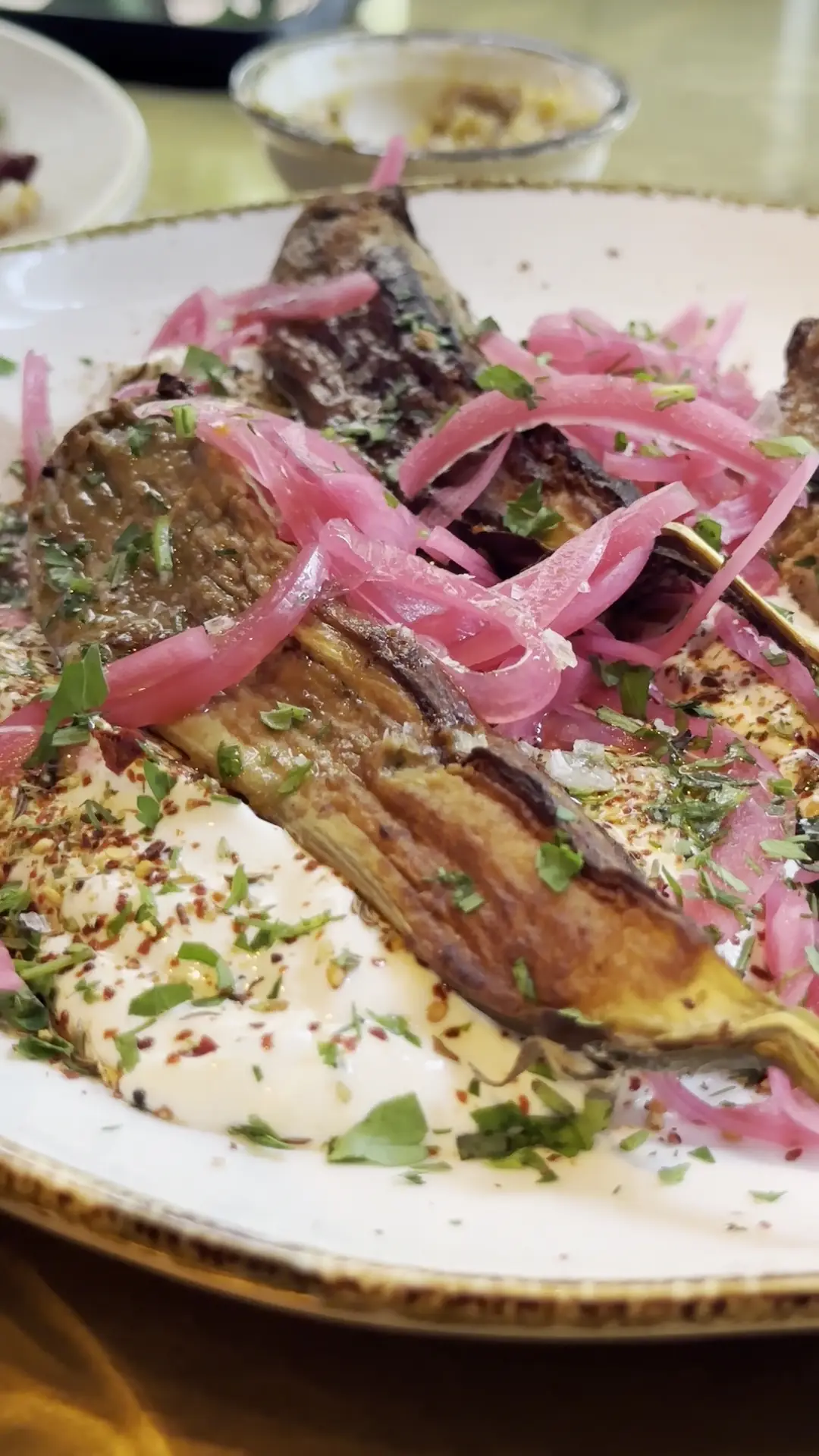 Grilled eggplant with pickled red onions over a white sauce, sprinkled with herbs.