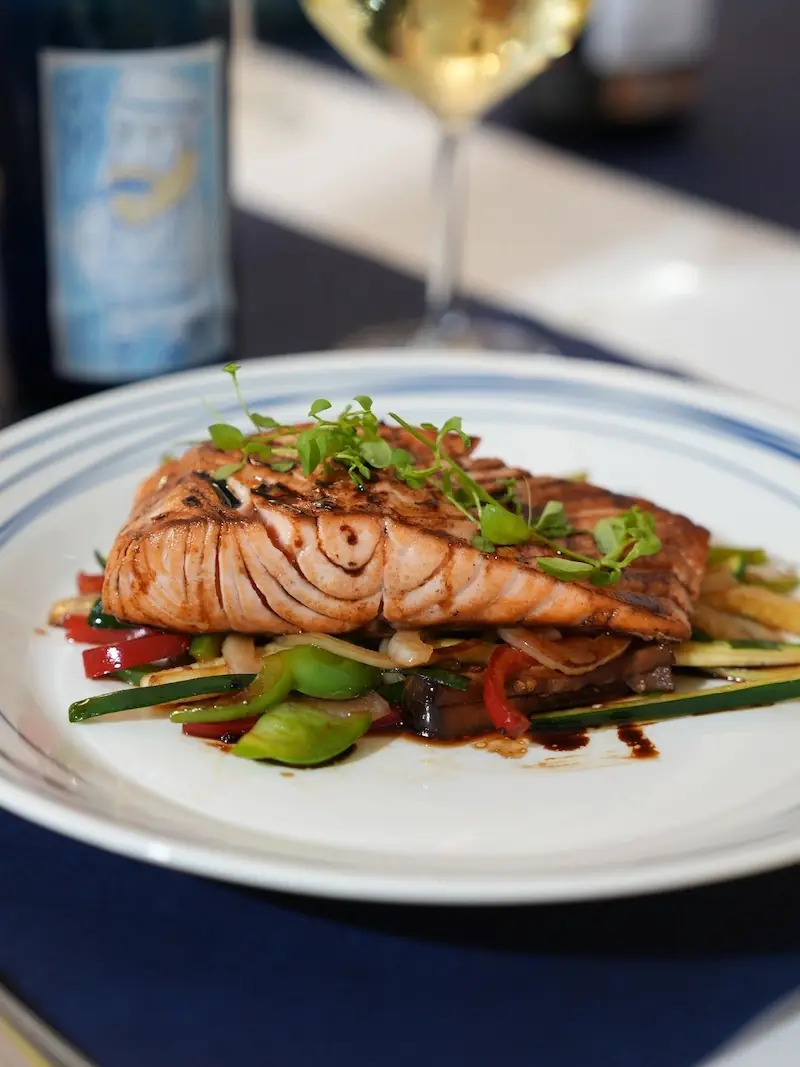 Grilled salmon fillet with charred grill marks and balsamic glaze, topped with microgreens, served over sautéed peppers, zucchini, and onions at A'Rogueira