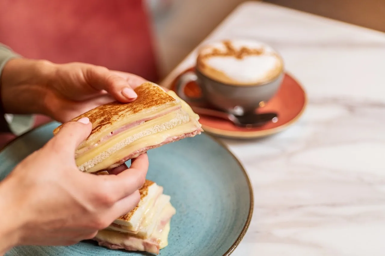 Hands holding a grilled sandwich next to a cappuccino