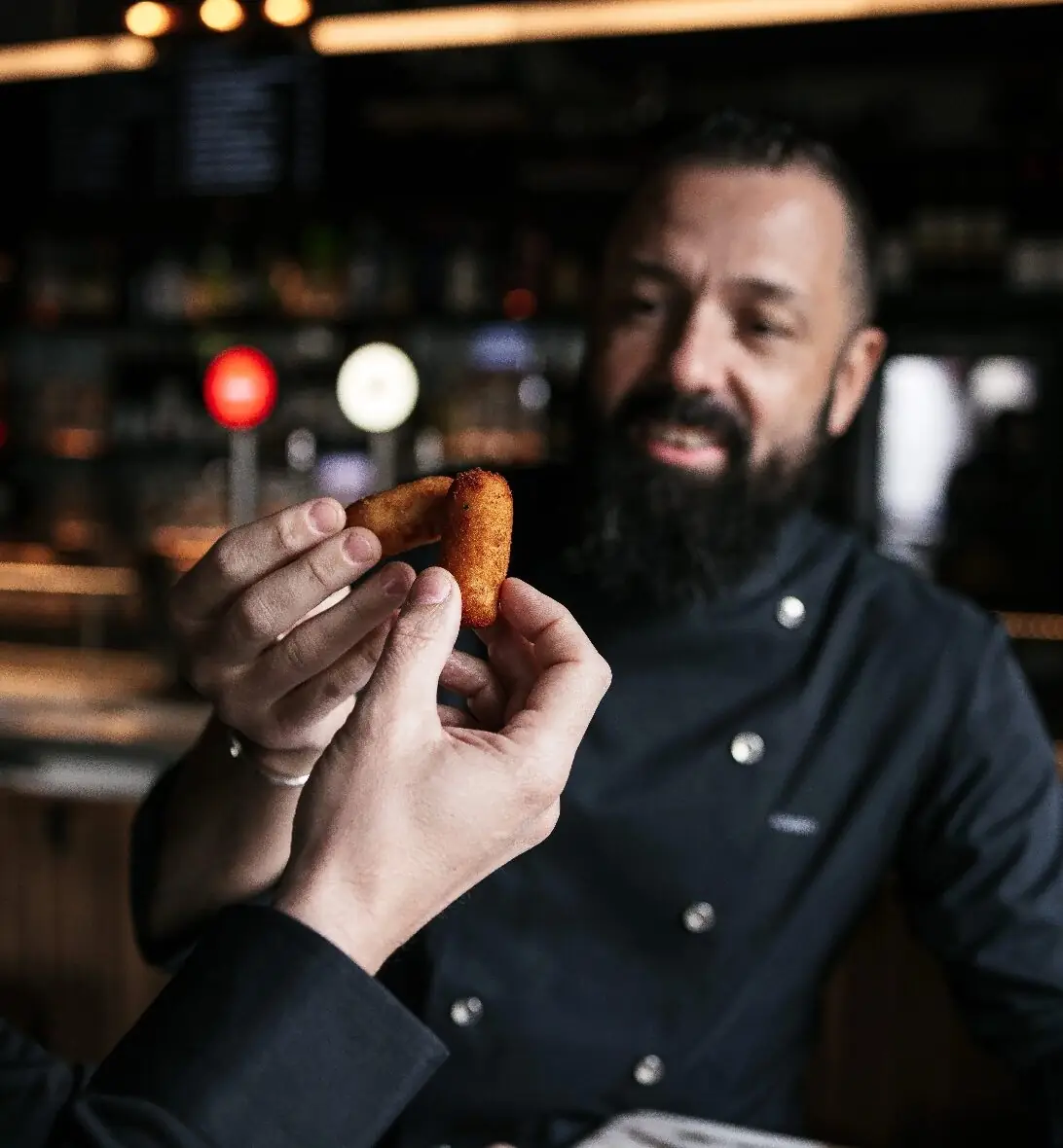 Two hands hold a single golden fried croqueta up to the camera in front of a dimly lit bar, with a bearded man in a dark chef's jacket blurred behind and beer taps and bottles out of focus in the background