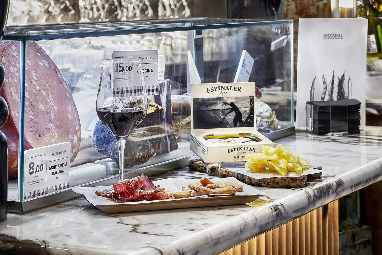 Glass of red wine beside a wooden board of jamon, breadsticks, chips and tinned sardines on a marble counter