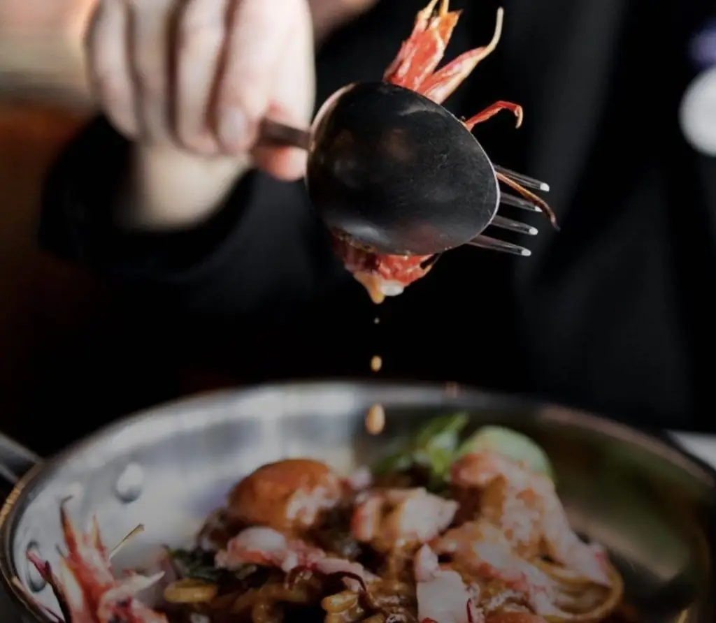 Hand spooning sauce over a prawn dish in a pan with greens