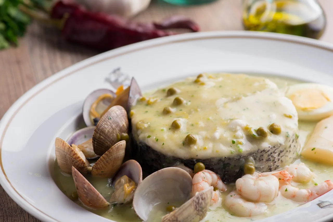 A thick fish steak in green sauce with peas, clams, prawns and a boiled egg on a white plate